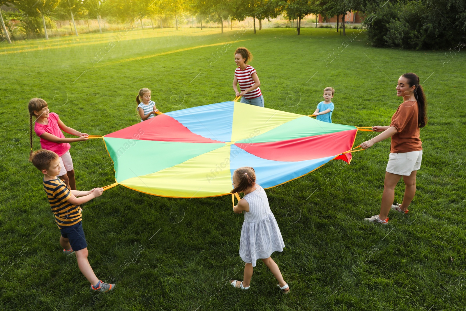 Photo of Group of children and teachers playing with rainbow playground parachute on green grass. Summer camp activity