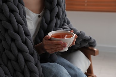 Photo of Woman with chunky knit blanket and cup of tea in armchair at home, closeup