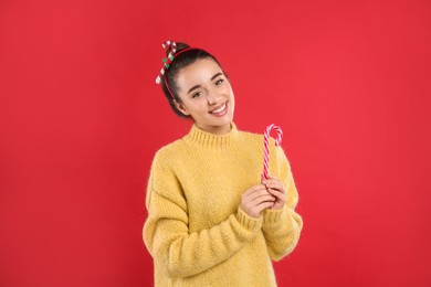 Photo of Young woman in yellow sweater and festive headband holding candy cane on red background