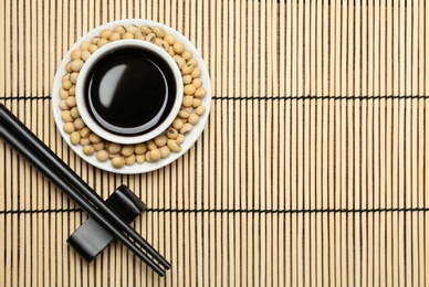 Photo of Dish of soy sauce with beans and chopsticks served on bamboo mat, flat lay. Space for text