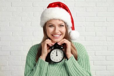 Photo of Happy young woman in Santa hat with alarm clock near white brick wall. Christmas time