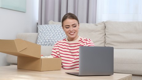 Photo of Happy woman with laptop unpacking parcel at home. Online store