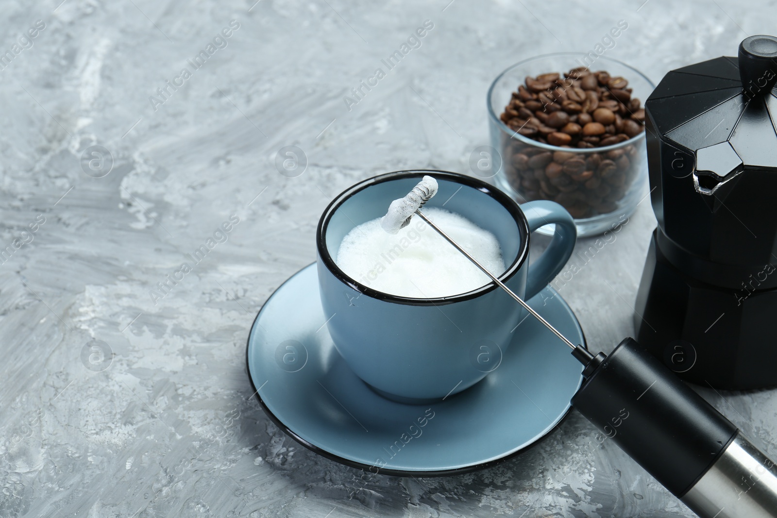 Photo of Mini mixer (milk frother), whipped milk in cup and coffee beans on grey textured table, closeup. Space for text