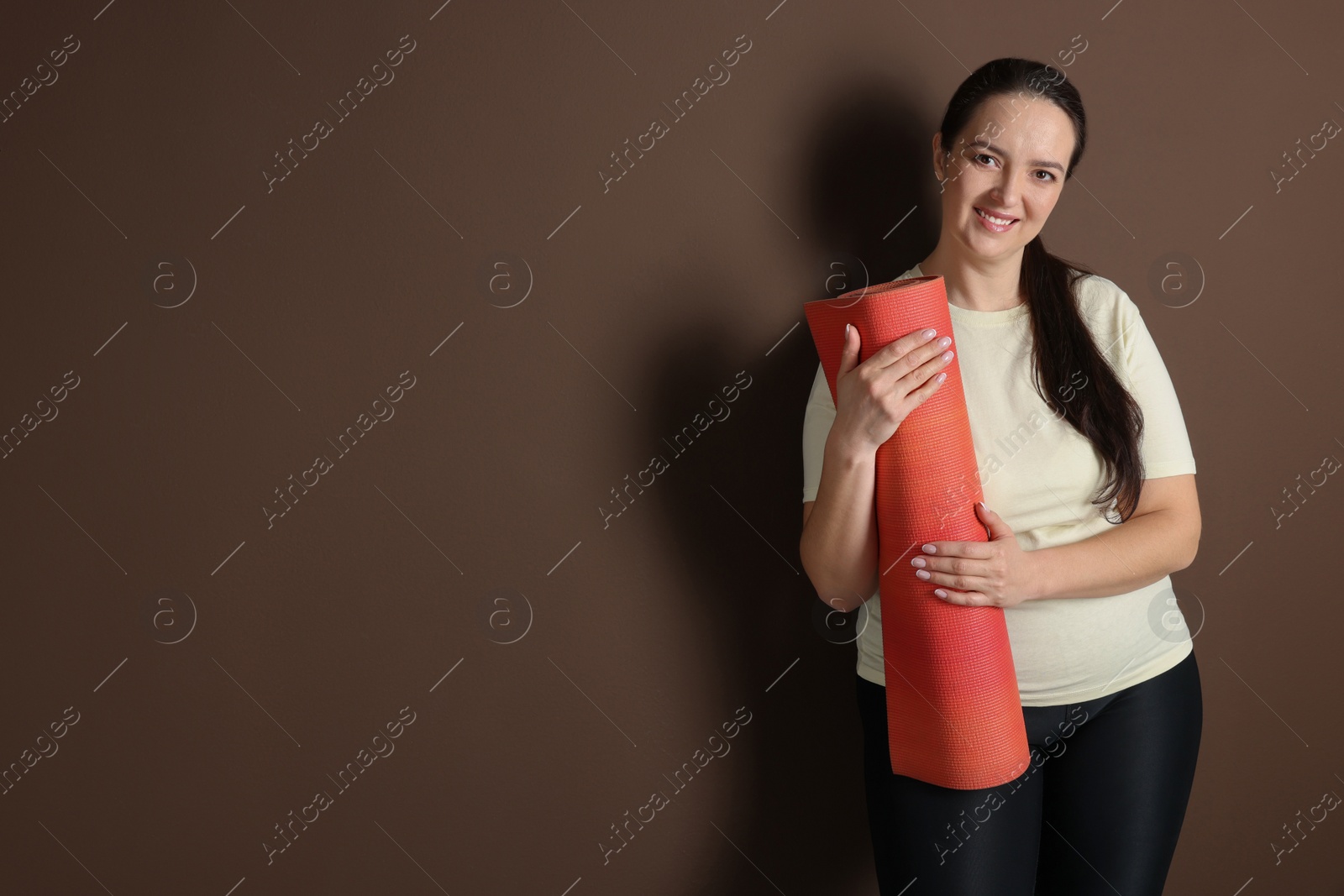 Photo of Happy overweight woman with yoga mat on brown background, space for text