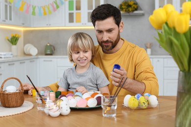 Photo of Father and his cute son painting Easter eggs at table in kitchen