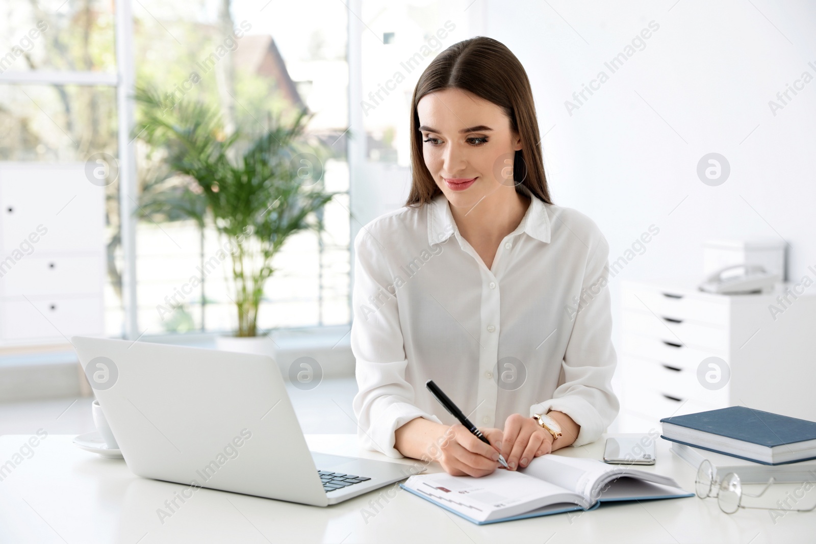 Photo of Young businesswoman writing in notebook while using laptop at table in office