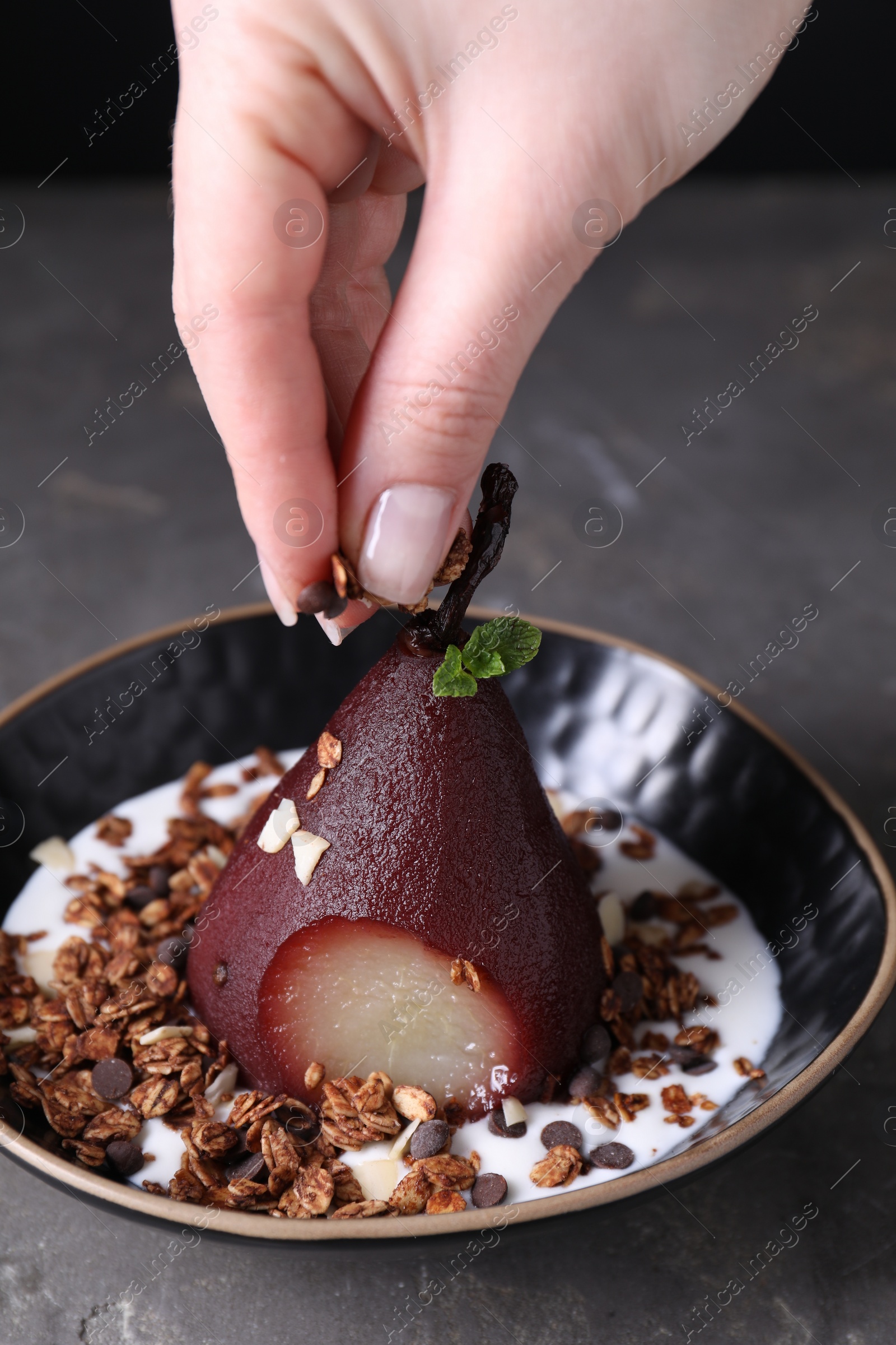 Photo of Woman putting muesli on tasty red wine poached pear in bowl at grey table, closeup