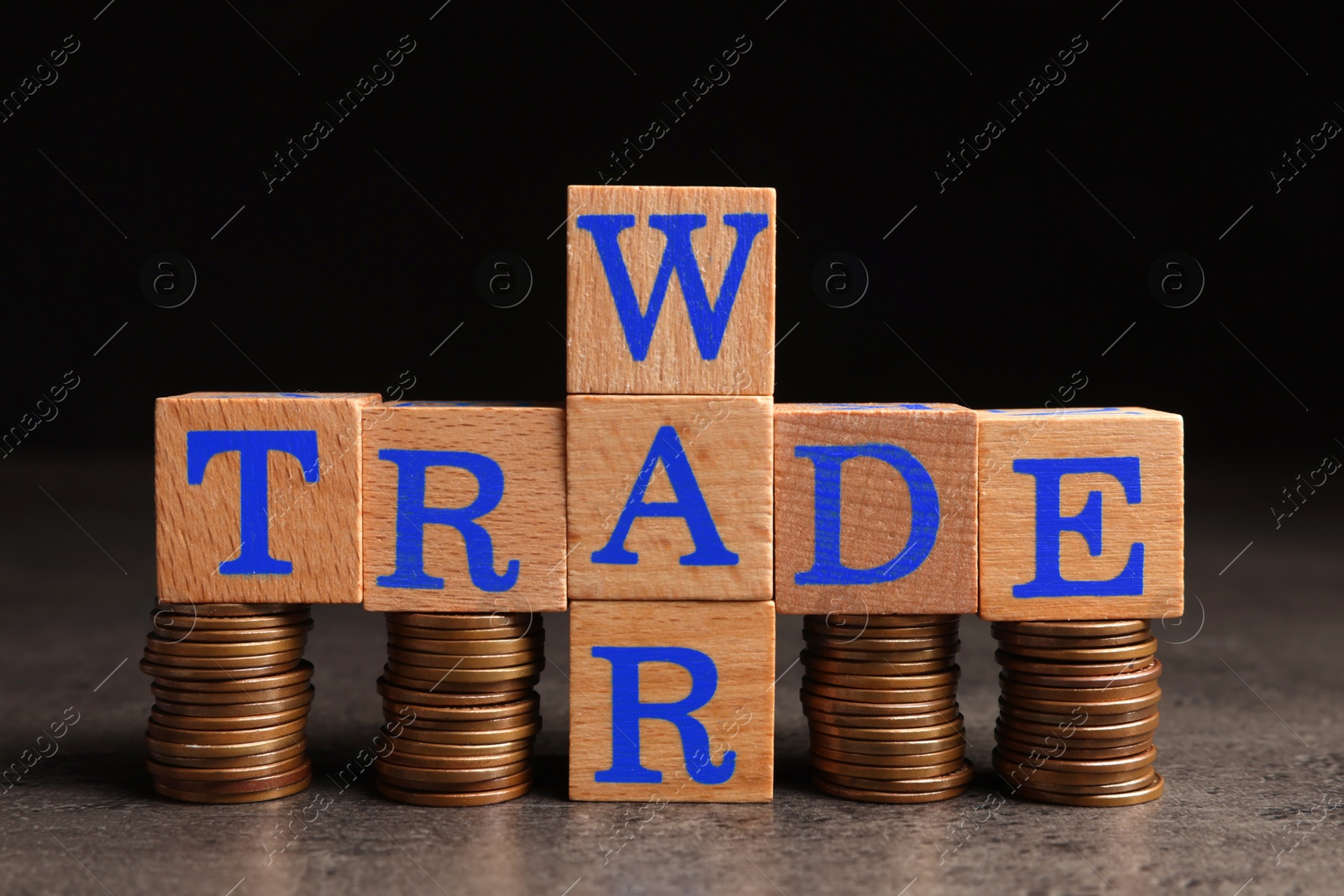 Photo of Words Trade war made of wooden cubes and stacked coins on grey table against black background, closeup