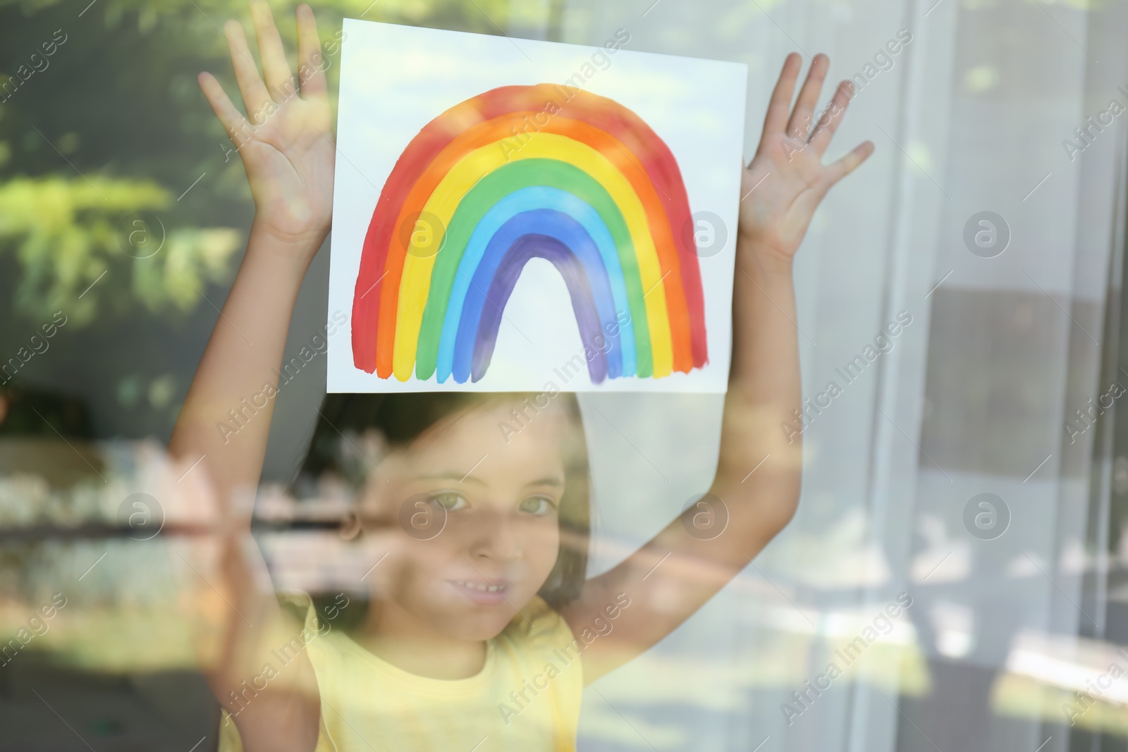 Photo of Little girl with picture of rainbow near window, view from outdoors. Stay at home concept