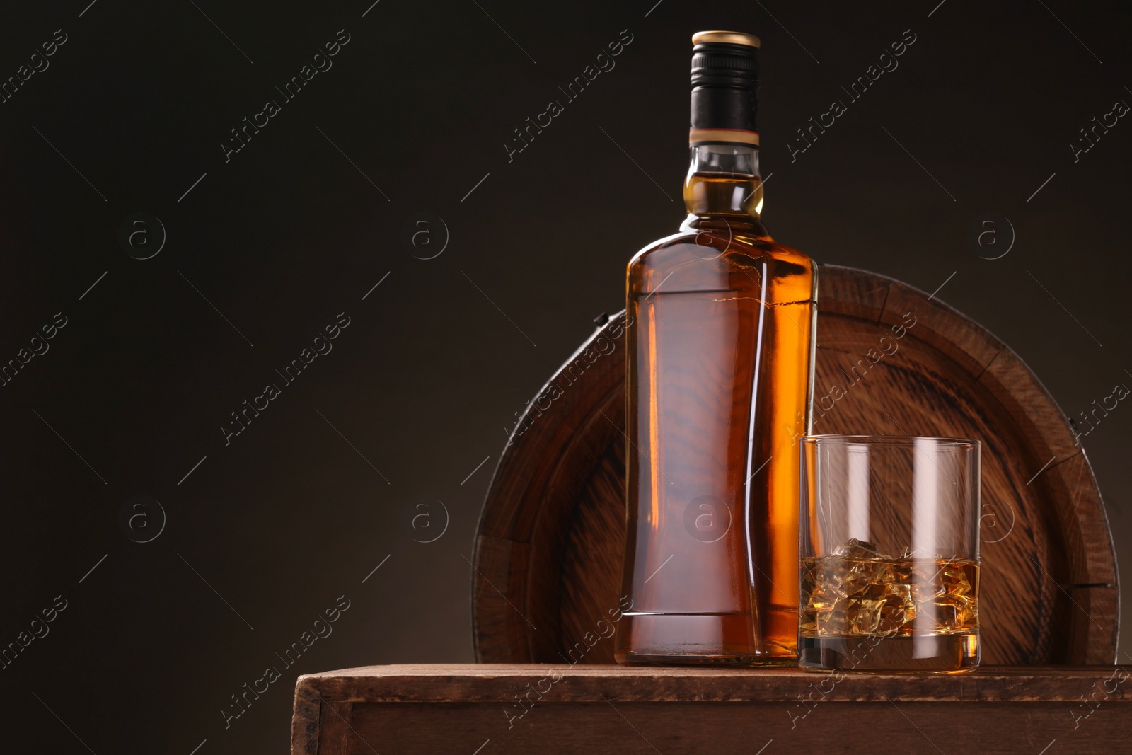 Photo of Whiskey with ice cubes in glass and bottle on wooden table near barrel against dark background, space for text