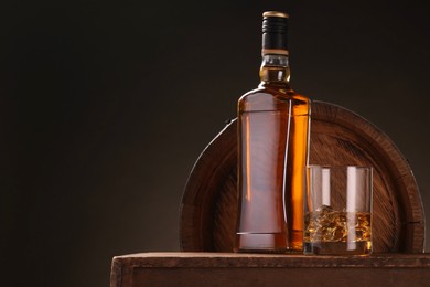 Whiskey with ice cubes in glass and bottle on wooden table near barrel against dark background, space for text