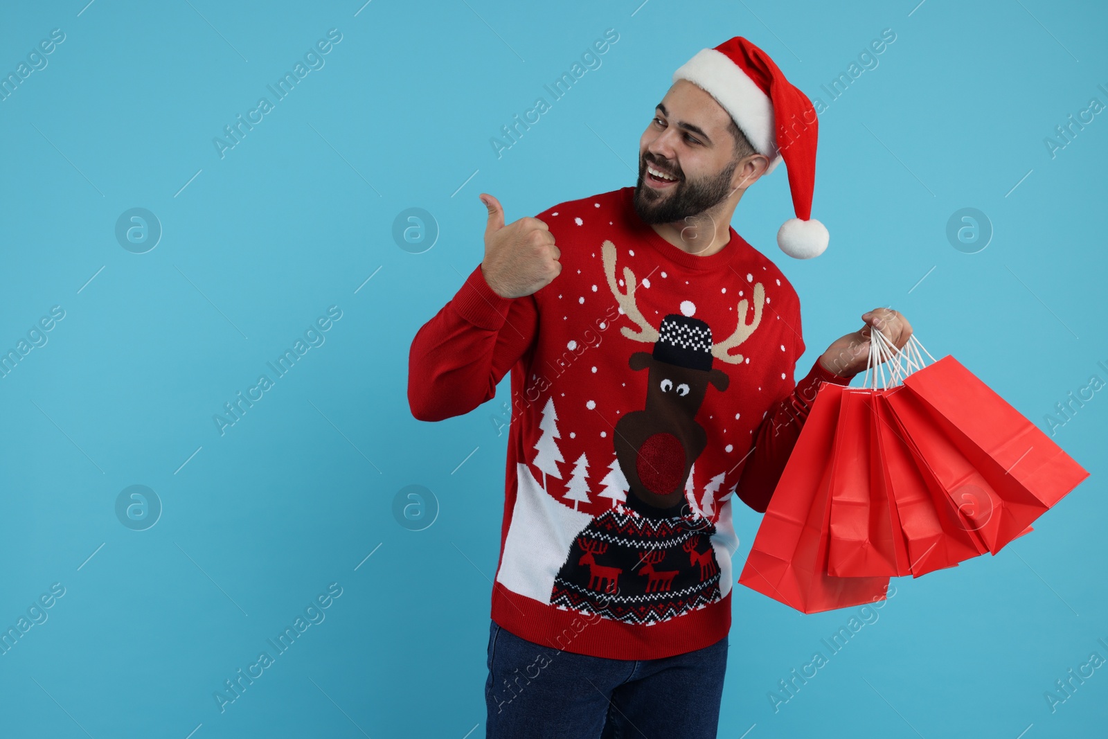 Photo of Happy young man in Christmas sweater and Santa hat with shopping bags on light blue background. Space for text