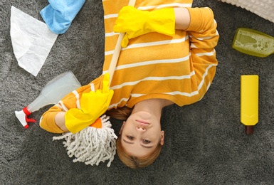 Photo of Lazy young woman with cleaning equipment lying on floor at home, top view