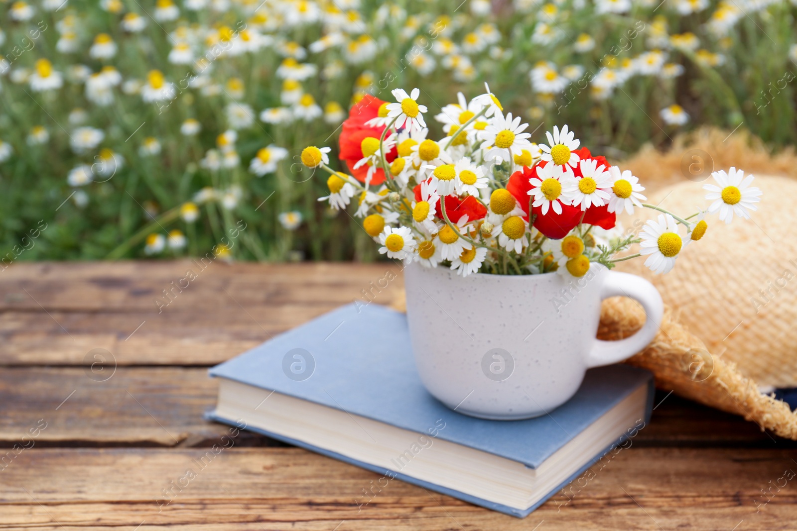 Photo of Composition with chamomiles, poppies, straw hat and book on wooden table outdoors