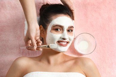 Cosmetologist applying white mask onto woman's face in spa salon, top view