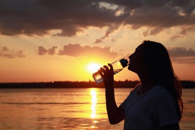 Photo of Young woman drinking water to prevent heat stroke near river at sunset, space for text