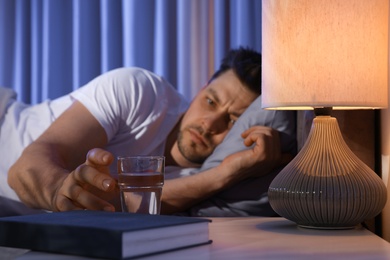 Photo of Sleepy man reaching for glass of water in dark room at night. Bedtime