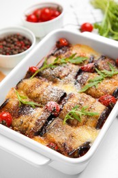 Photo of Tasty eggplant rolls with tomatoes, cheese and arugula in baking dish on white table, closeup