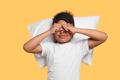 Photo of Boy with pillow and sleep mask rubbing his eyes on yellow background. Insomnia problem