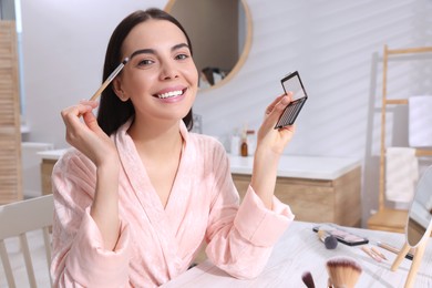 Beautiful young woman applying eyeshadow with brush at dressing table indoors