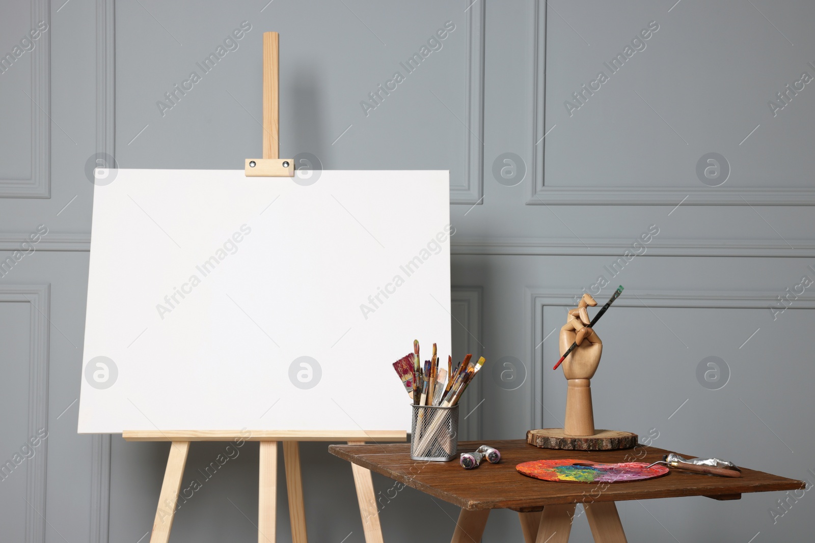 Photo of Easel with blank canvas and different art supplies on wooden table near grey wall