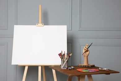 Photo of Easel with blank canvas and different art supplies on wooden table near grey wall