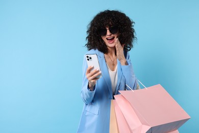 Photo of Happy young woman with shopping bags and modern smartphone on light blue background. Space for text