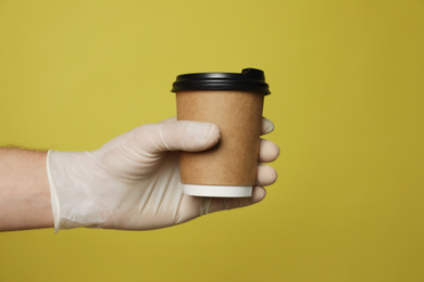 Photo of Courier in protective gloves holding cup with drink on yellow background, closeup of hand. Delivery service during coronavirus quarantine