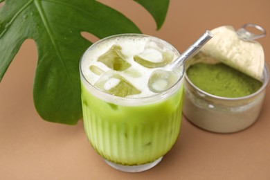 Glass of tasty iced matcha latte, powder and leaf on light brown background, closeup