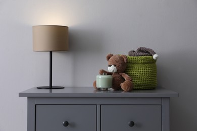 Photo of Lamp, child's toy and wicker basket on chest of drawers near light grey wall indoors