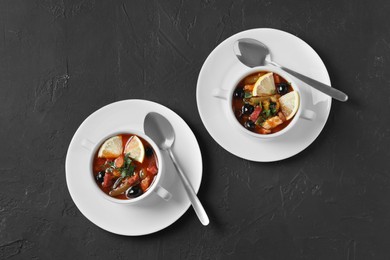 Photo of Meat solyanka soup with sausages, olives, vegetables and spoons on dark grey textured table, flat lay