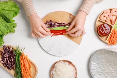 Photo of Making delicious spring rolls. Woman wrapping fresh vegetables into rice paper at white wooden table, top view