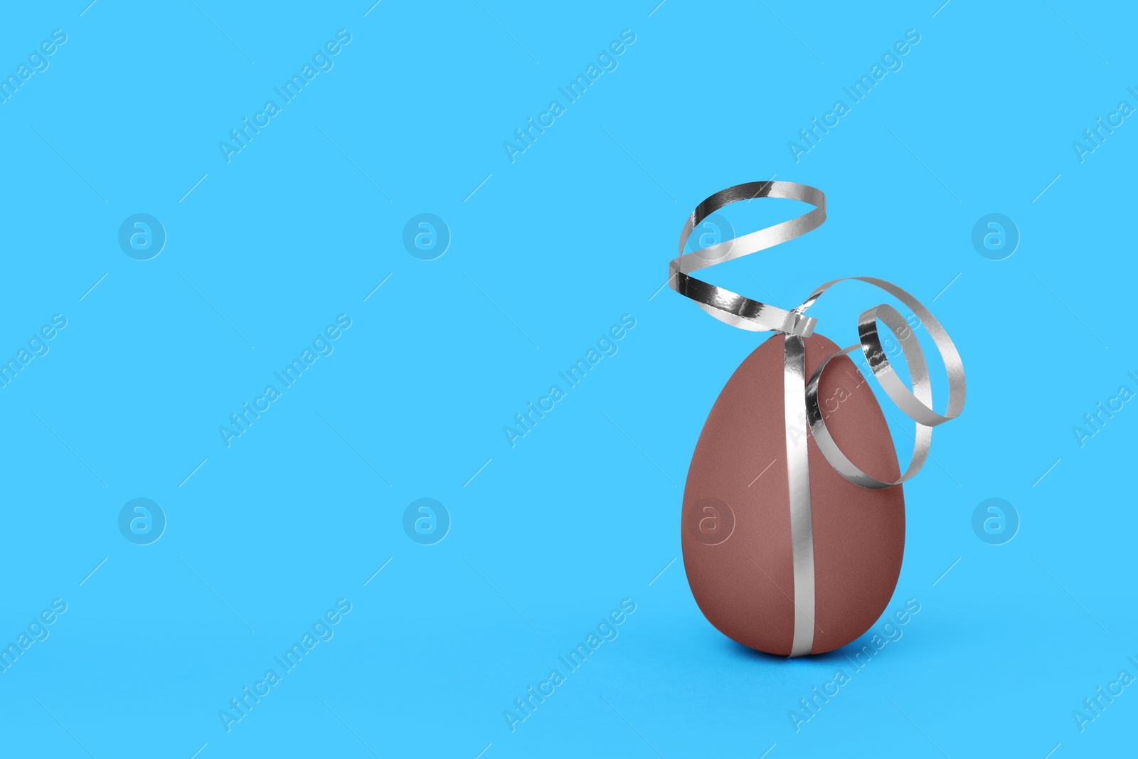Delicious chocolate egg with silver ribbon on light blue background. Space for text Photo of Delicious chocolate egg with silver ribbon on light blue background. Space for text