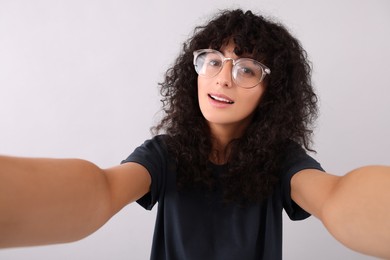 Beautiful young woman in eyeglasses taking selfie on light grey background