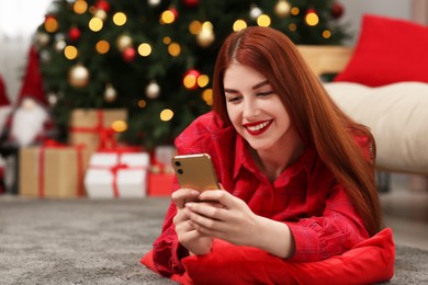 Photo of Beautiful young woman using smartphone near Christmas tree at home, space for text