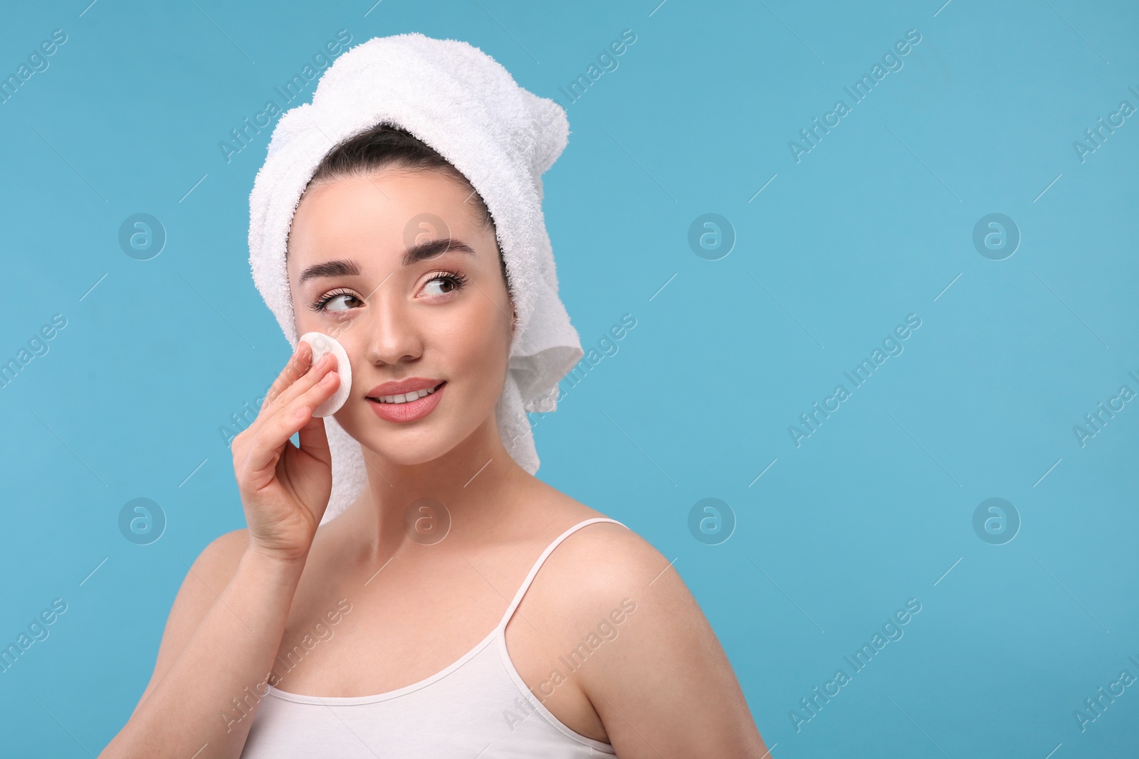 Photo of Beautiful woman in terry towel removing makeup with cotton pad on light blue background, space for text