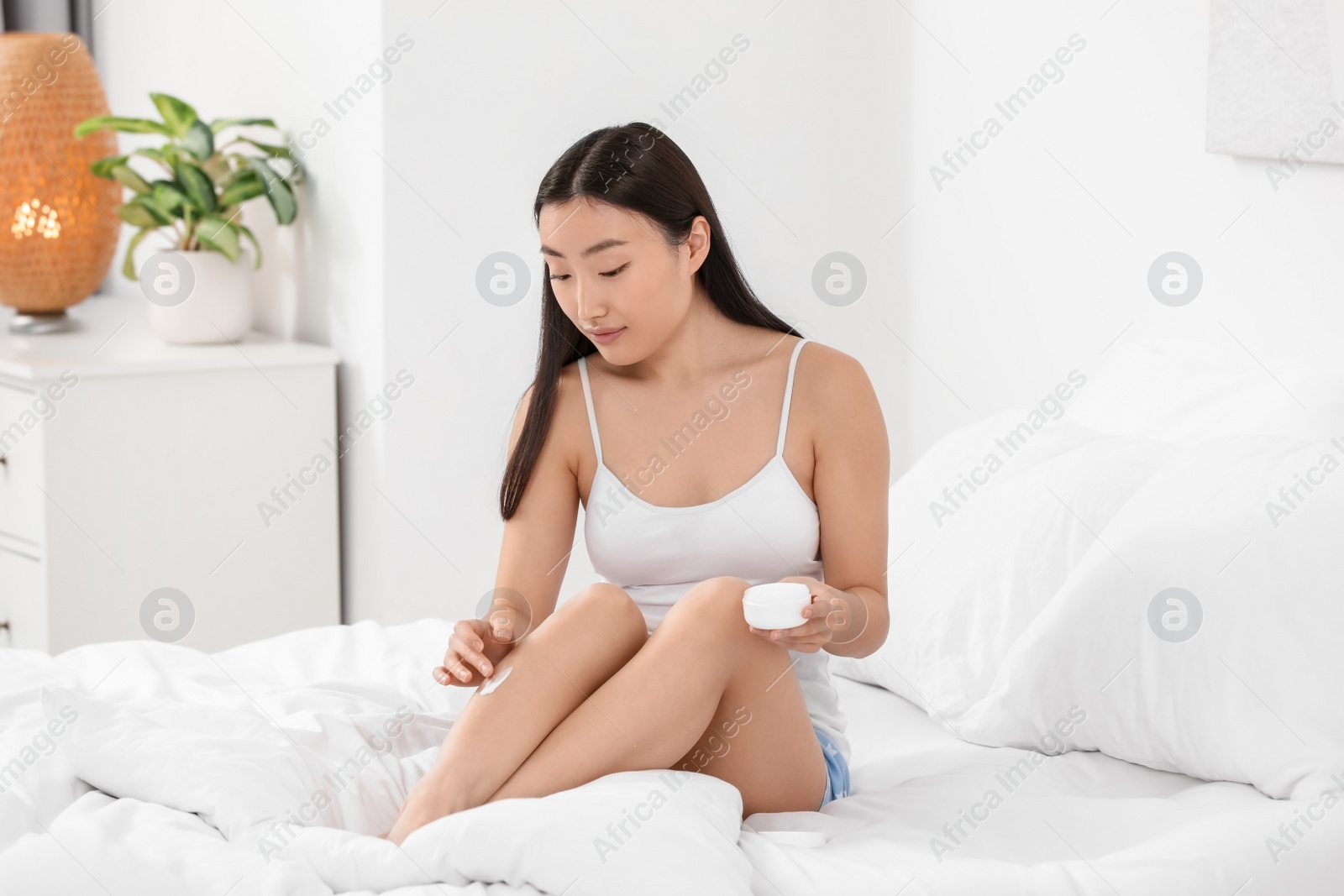 Photo of Beautiful young Asian woman applying body cream on leg in bedroom