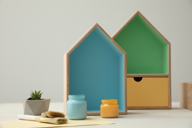 Photo of House shaped shelves, jars of paints and brushes on white table. Interior elements