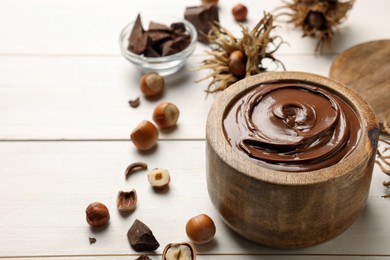 Photo of Bowl of tasty chocolate paste with hazelnuts on white wooden table, space for text