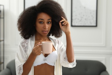 Beautiful woman in elegant white underwear and robe holding cup of drink indoors, space for text
