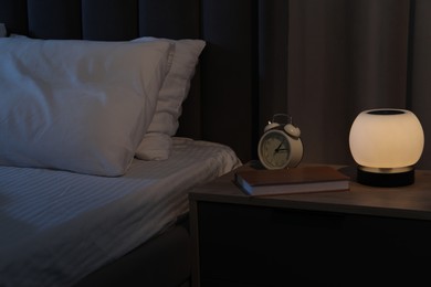 Photo of Nightlight, alarm clock and book on bedside table near bed indoors