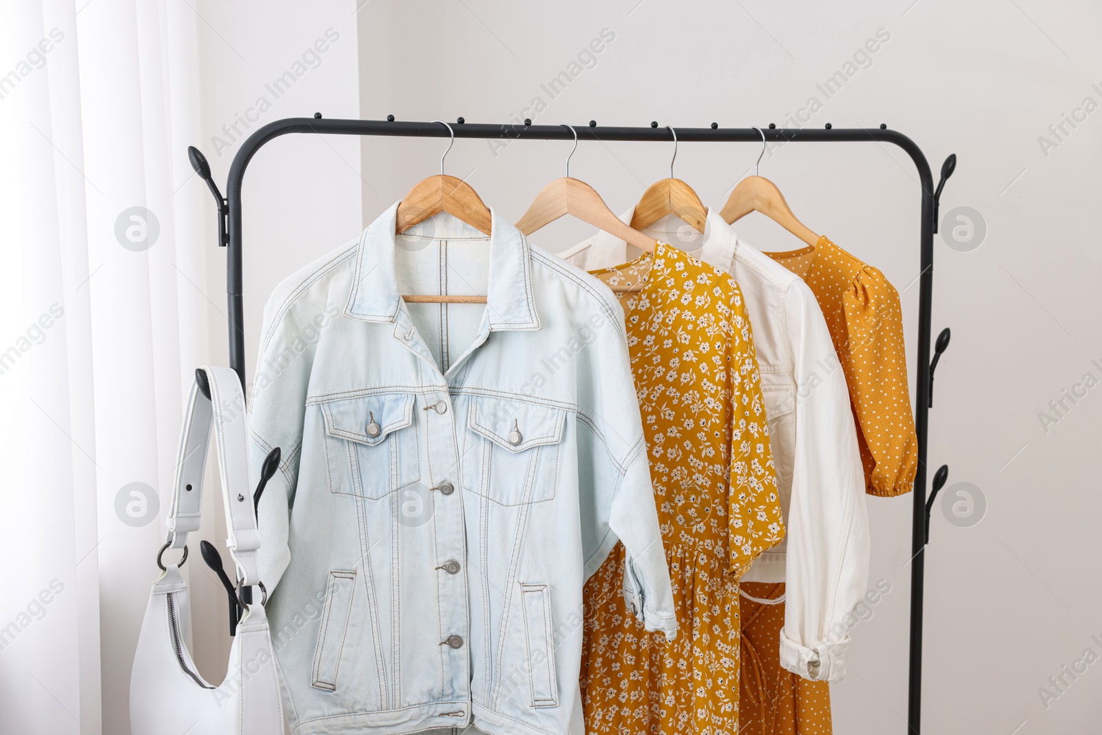 Photo of Rack with bag and stylish women's clothes on hangers near light wall indoors