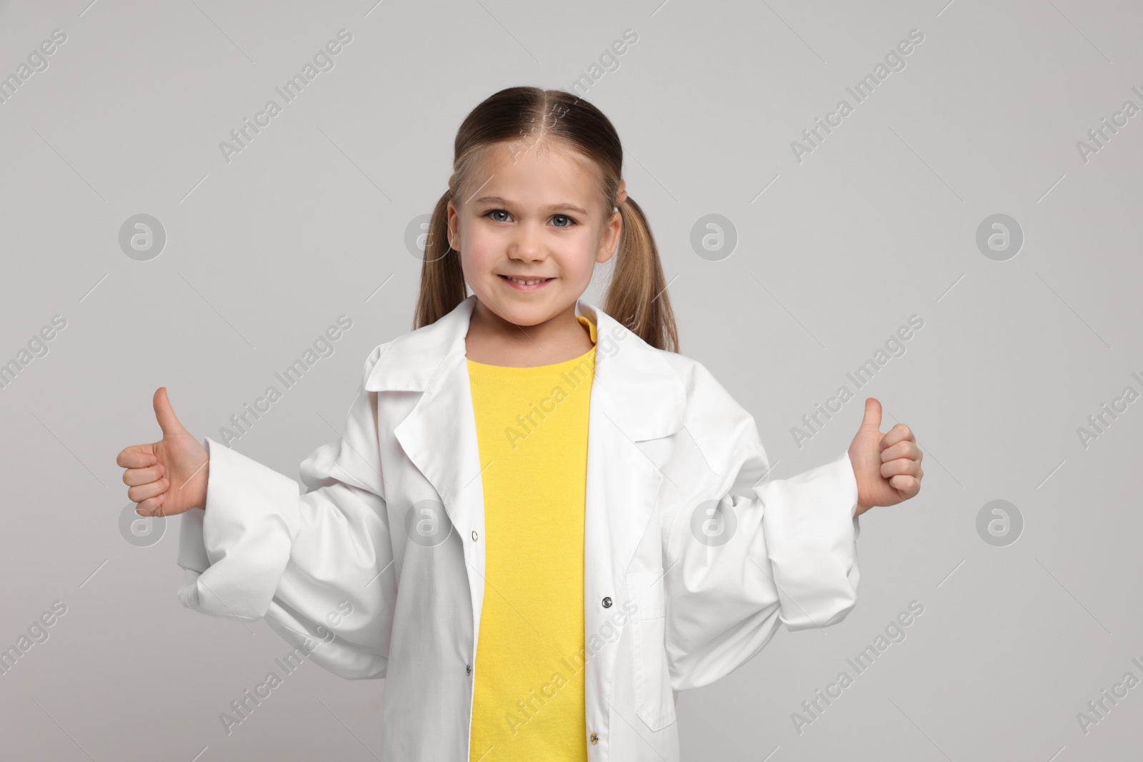 Photo of Little girl in medical uniform showing thumbs up on light grey background