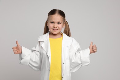Photo of Little girl in medical uniform showing thumbs up on light grey background
