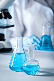 Photo of Closeup view of scientist pouring light blue liquid from test tube into flask and laboratory glassware on table, selective focus