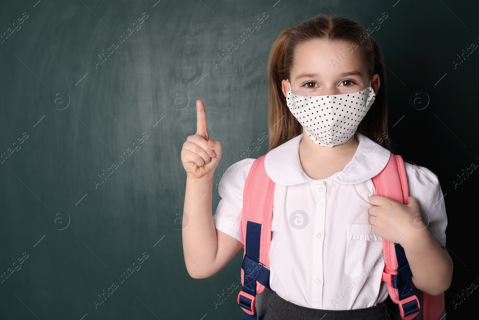 Photo of Little girl wearing protective mask and backpack near chalkboard, space for text. Child safety