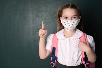 Photo of Little girl wearing protective mask and backpack near chalkboard, space for text. Child safety
