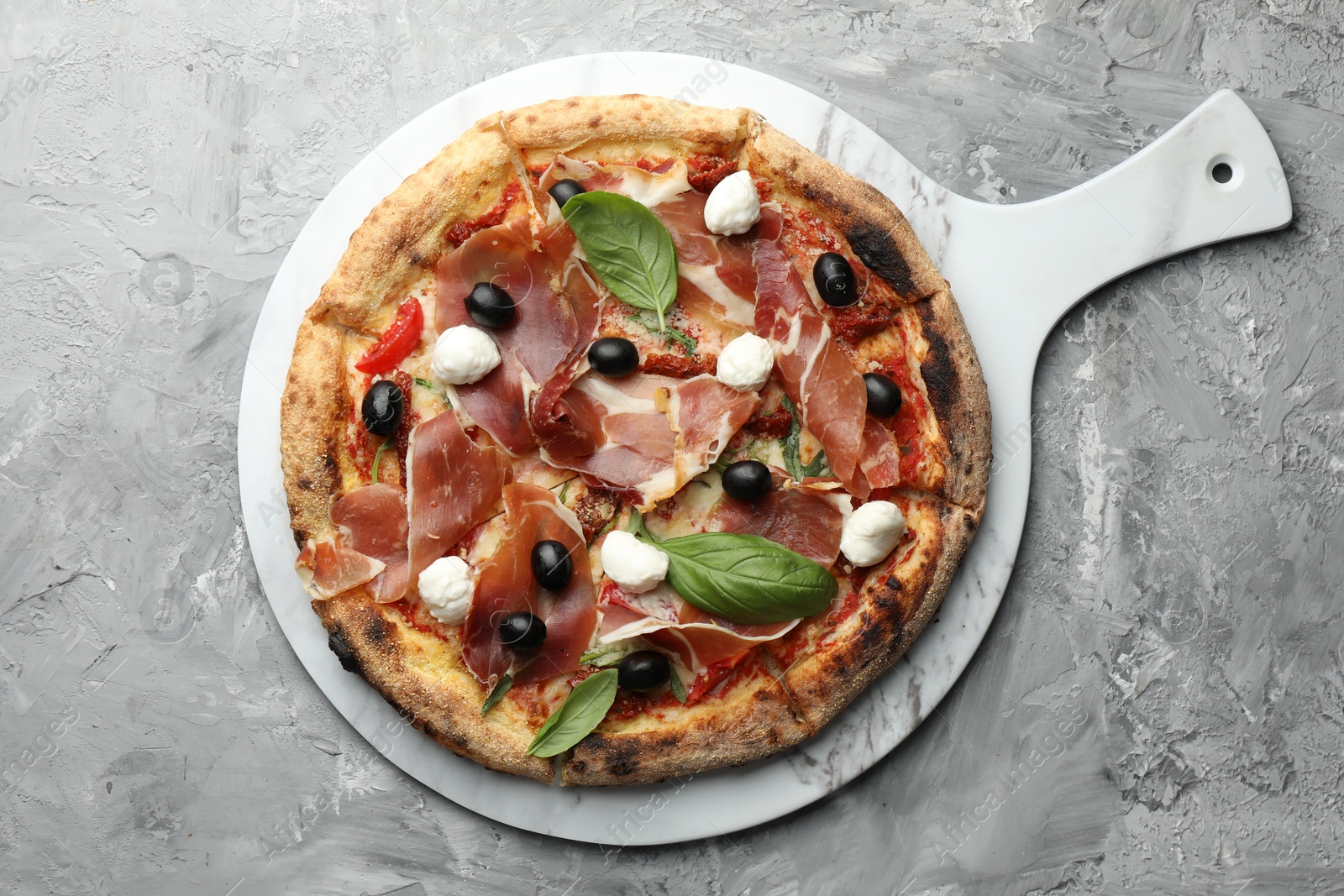 Photo of Tasty pizza with cured ham, olives, sun-dried tomato and basil on gray textured table, top view