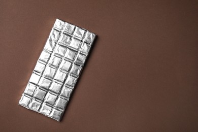 Photo of Tasty chocolate bar wrapped in foil on brown background, top view. Space for text