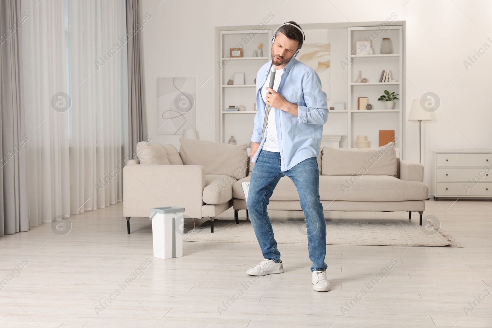 Photo of Enjoying cleaning. Happy man in headphones with mop singing while tidying up at home